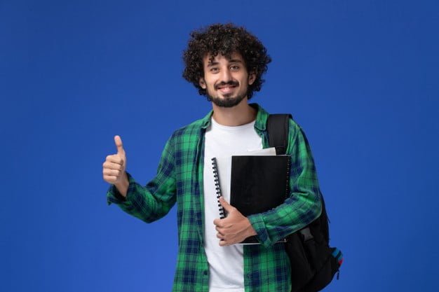 front view male student green checkered shirt with black backpack holding copybooks smiling blue wall 140725 42439